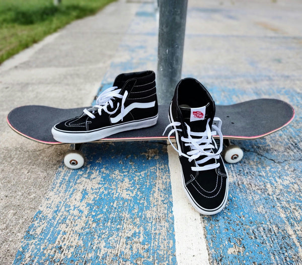 Pair of Vans skate shoes Sk8-Hi Black and White sneakers with white laces resting on a skateboard at a skatepark.