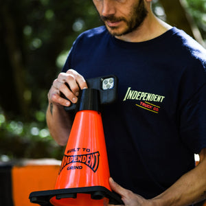 A man wearing a navy blue t-shirt with 'INDEPENDENT TRUCK CO' printed on the chest holds an orange traffic cone branded with 'BUILT TO INDEPENDENT GRIND' in an outdoor setting.