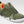 An olive green athletic sneaker with a white sole, featuring a textured mesh and suede upper, green laces, and a bright orange interior lining.