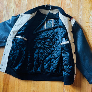 A vintage Husqvarna jacket laid flat on a wooden floor, featuring a dark blue quilted interior, black leather sleeves and collar, and beige ribbed knit trim.