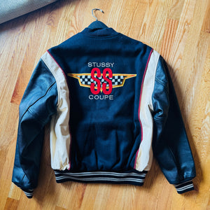 A vintage Stussy 88 Coupe leather jacket laid flat on a wooden floor, showing the back with cream leather sleeves, a navy body, and a large red and yellow number 88 graphic.