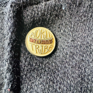 A round gold-tone metal pin with the words 'WORLD STUSSY TRIBE' in raised lettering, placed on a dark textured fabric background.
