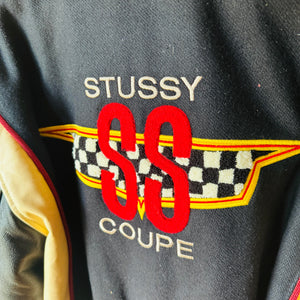 Close-up of a dark gray embroidered patch reading 'STUSSY SS COUPE' with red lettering, a checkered flag motif, and yellow trim on a garment.